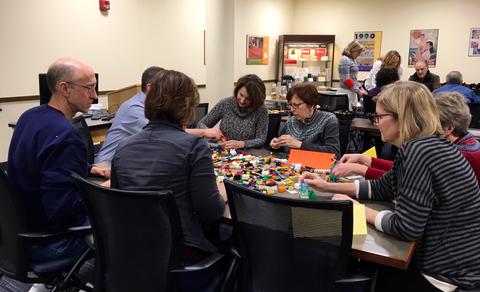 Photograph of a group of people sitting at a table building something out of building blocks.