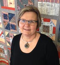 Photograph of Jane Peterson, upper torso and head standing in front of the MPC quilt.