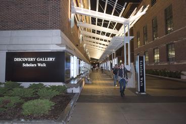 Students walk along outdoor sidewalk labelled "Discovery Gallery, Scholars Walk"
